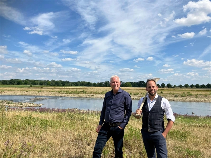 Links Hermjan Barneveld en rechts Roy Frings, staand in de uiterwaarden met gras en water en een blauwe lucht met witte wolken.