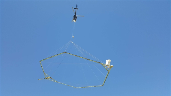 Foto met helicopter tegen blauwe lucht. Onder de helicopter hangt een groot 6-hoekig raamwerk aan touwen.