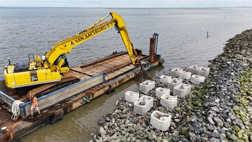 De foto toont een ponton in het water met daarop een graafmachine. Op de stenige kust staan grote witte bakken.