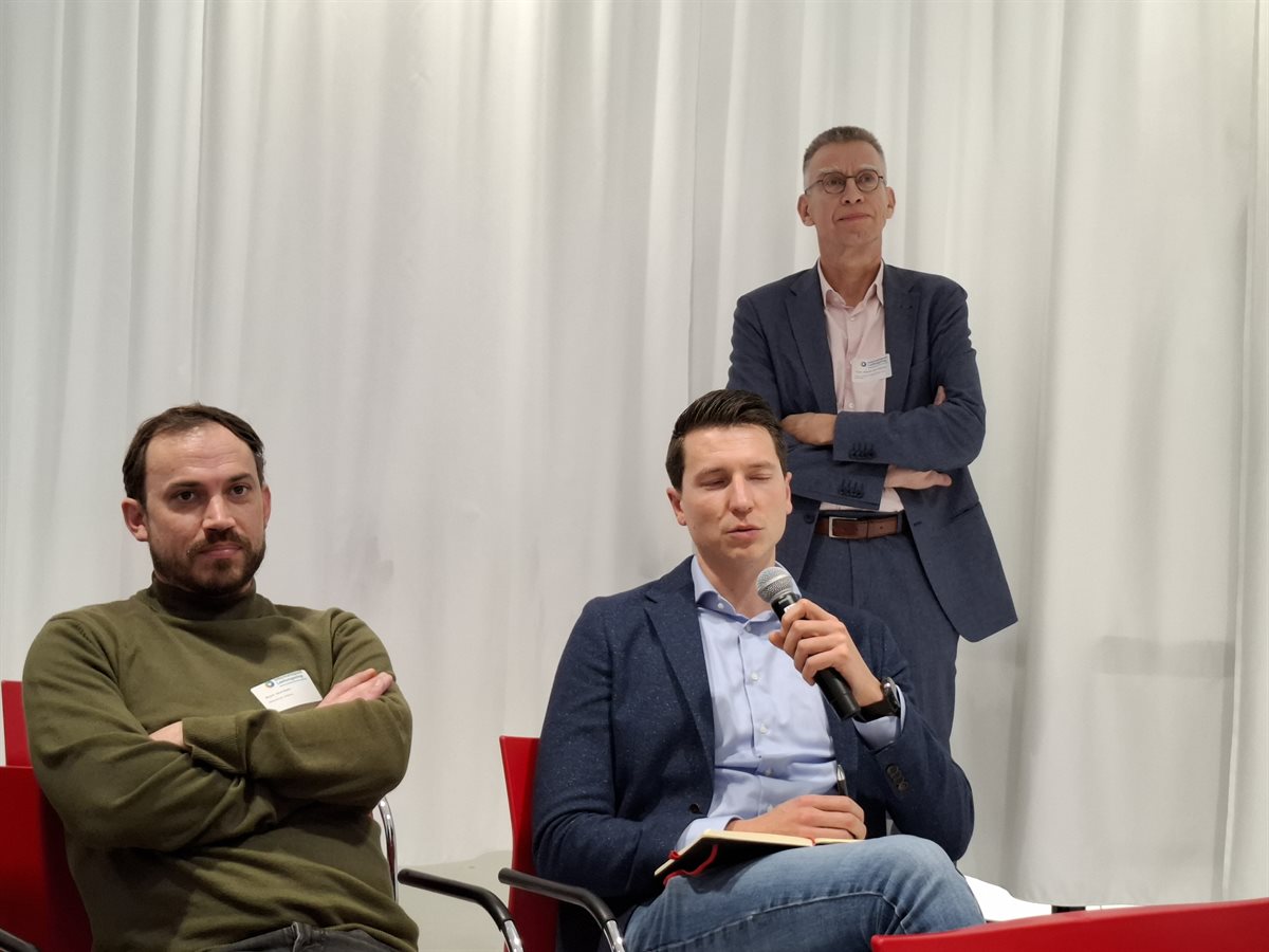 Drie deelnemers, van wie er een de microfoon in zijn hand heeft.